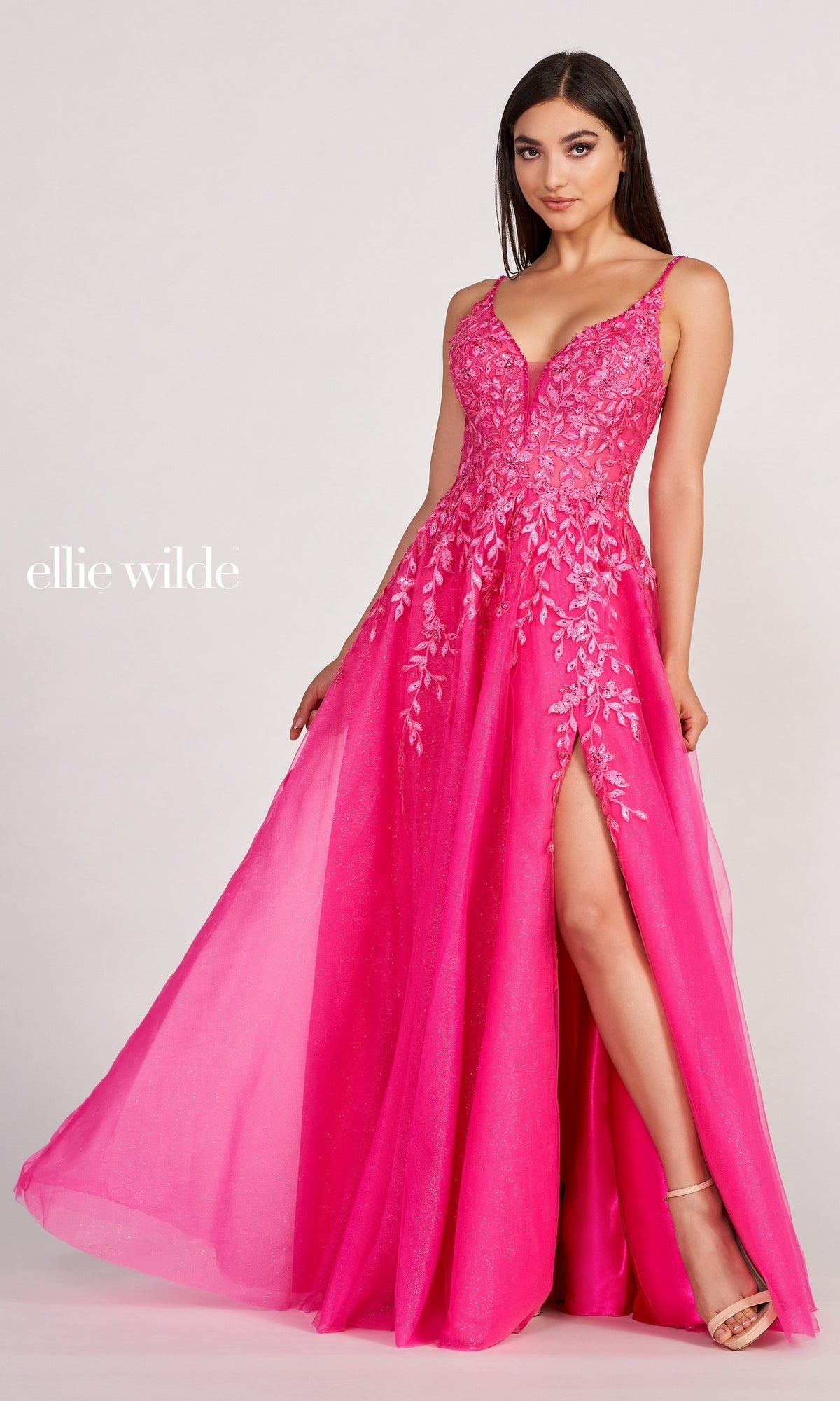 A woman poses against a plain background wearing the Glitter-Tulle Long Ellie Wilde Prom Dress EW34089, a bright pink, floor-length gown with floral embellishments, crystal beading, a thigh-high slit, and spaghetti straps.